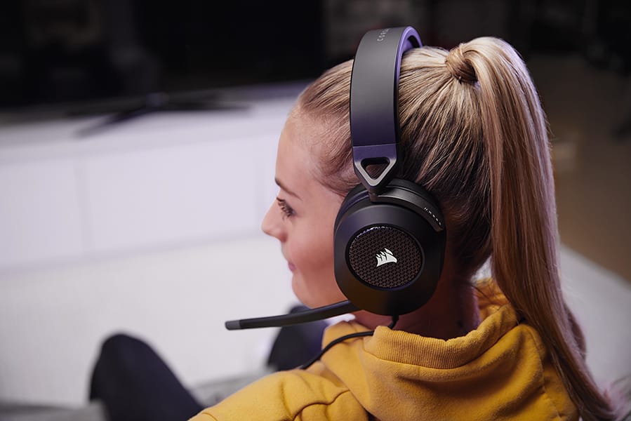 A pair of black HS65 SURROUND wired gaming headset on a woman, facing sideways, sitting on a couch.