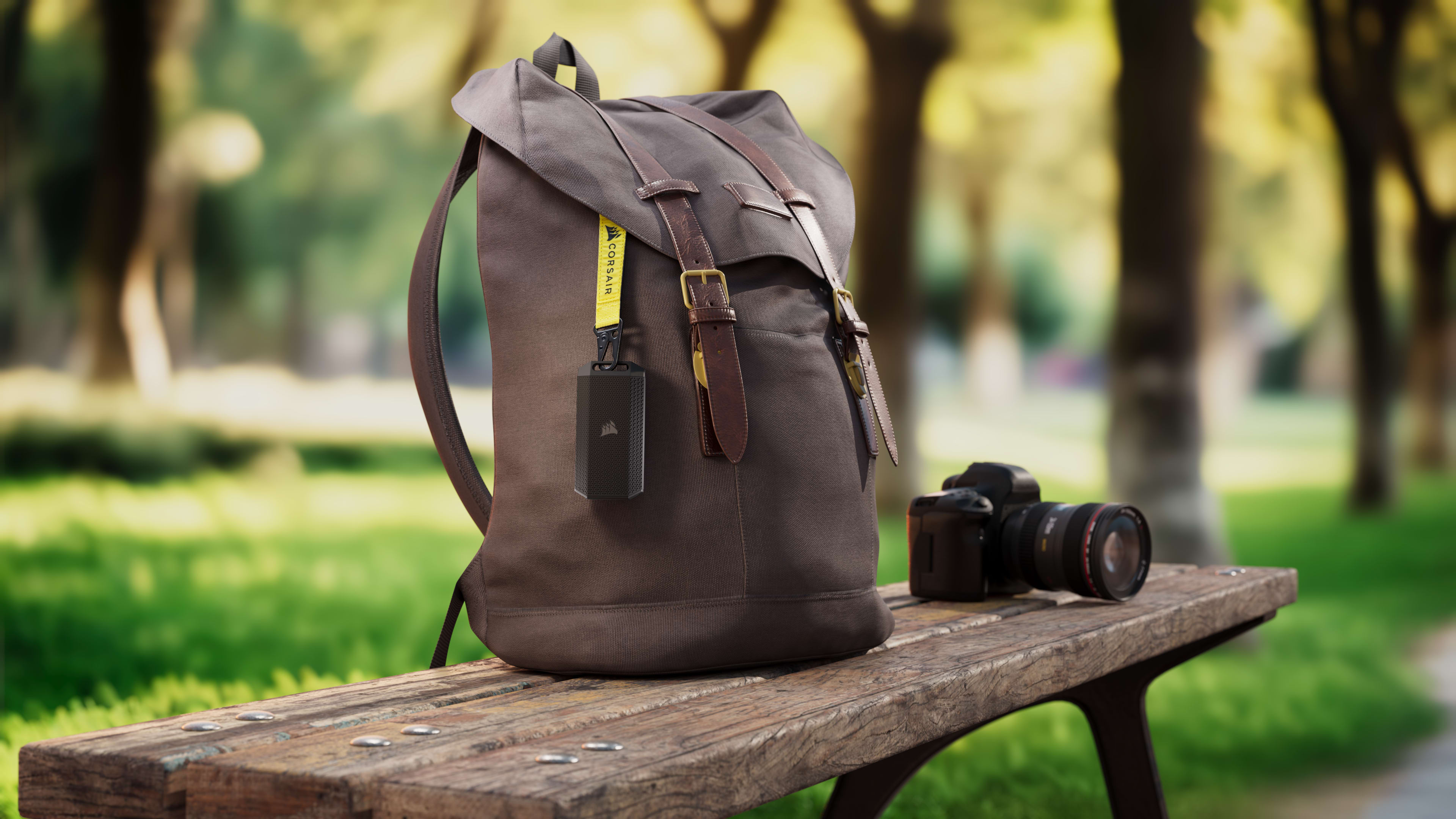 CORSAIR EX400U Survivor portable SSD clipped to a backpack strap on a park bench alongside a DSLR camera, illustrating on-the-go storage reliability.