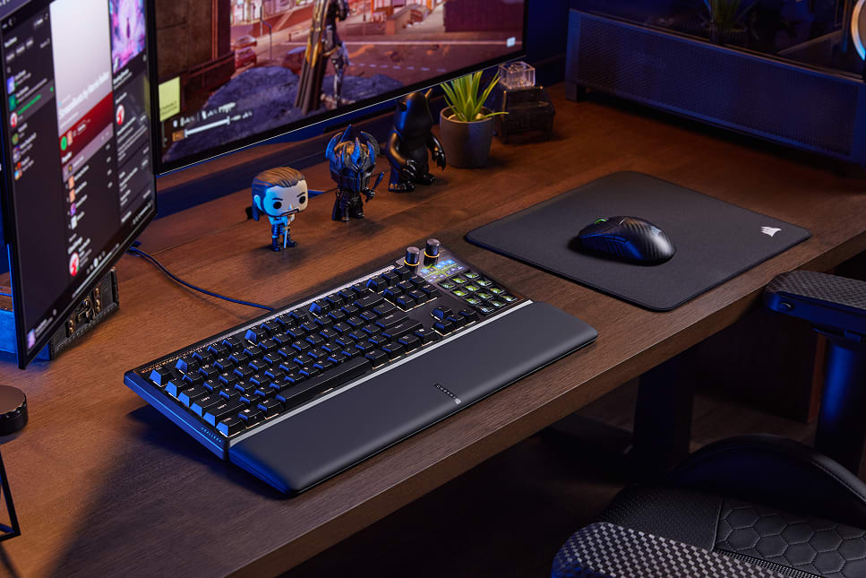 A gaming desk setup featuring a Corsair keyboard with blue LED backlighting, a mouse, monitor, and decorative figurines with ambient purple lightings