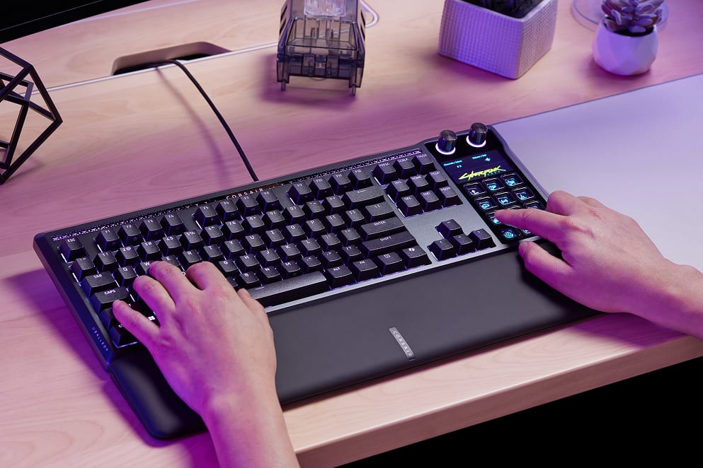 Close-up of two hands typing on an illuminated Corsair gaming keyboard with purple and pink backlighting on a desks