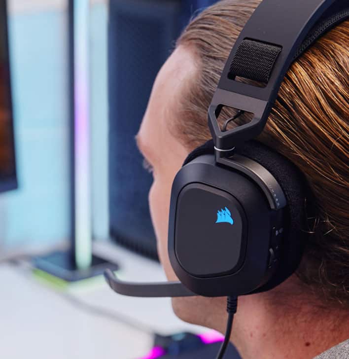 Man wearing HS80 RGB USB wired gaming headset while playing video games with RGB lighting on the Corsair sail logo.