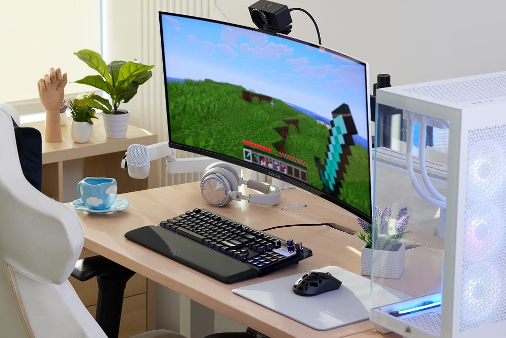A bright home office desk setup featuring a curved monitor displaying a scenic landscape, a Corsair keyboard, and a decorative plants