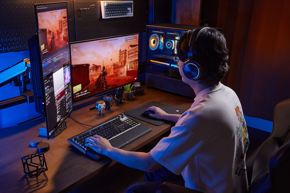 A person wearing a blue gaming headset seated at a multi-monitor gaming setup with blue ambient lighting and a Corsair keyboards