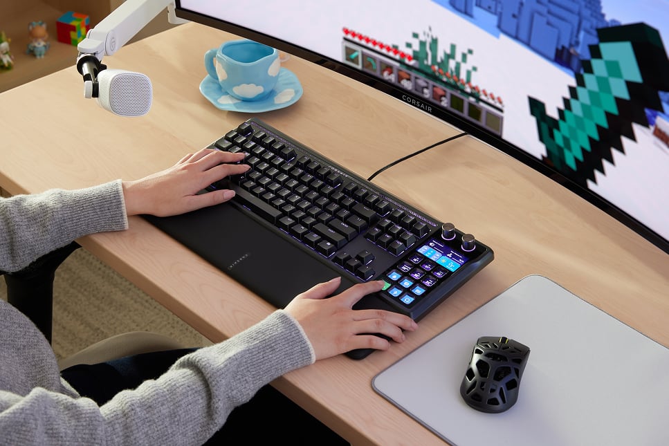 A person in a grey sweater typing on a Corsair keyboard at a bright wooden desk with a monitor and accessories
