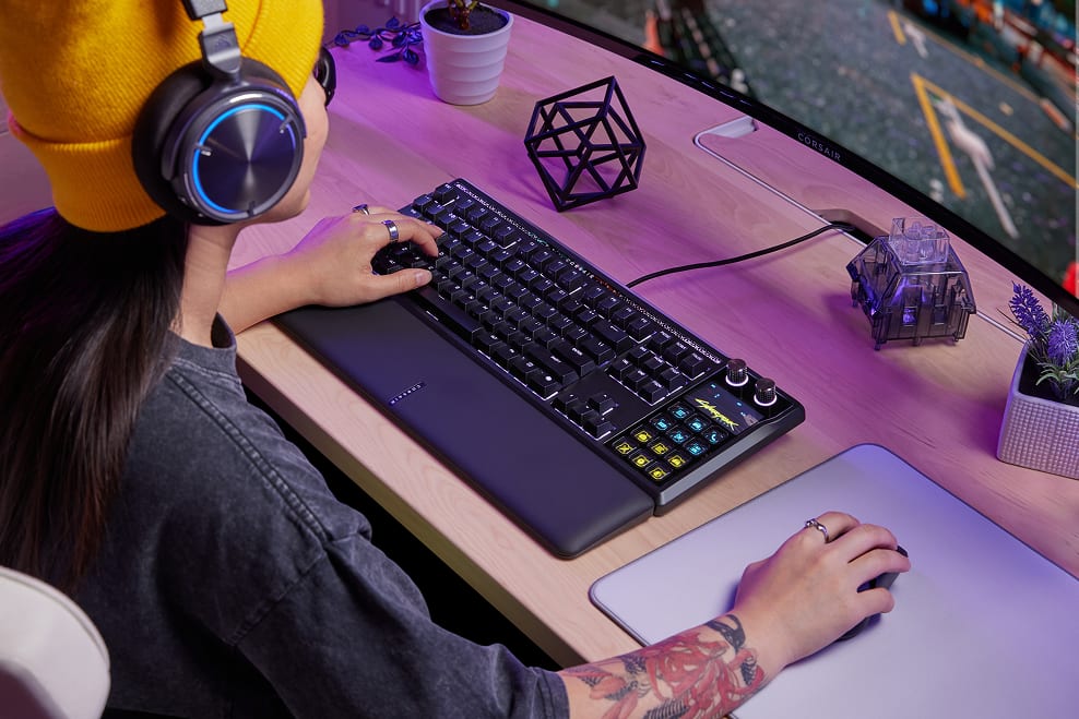 A person with tattoos and yellow headphones using a Corsair gaming keyboard at a purple-lit gaming desk setups