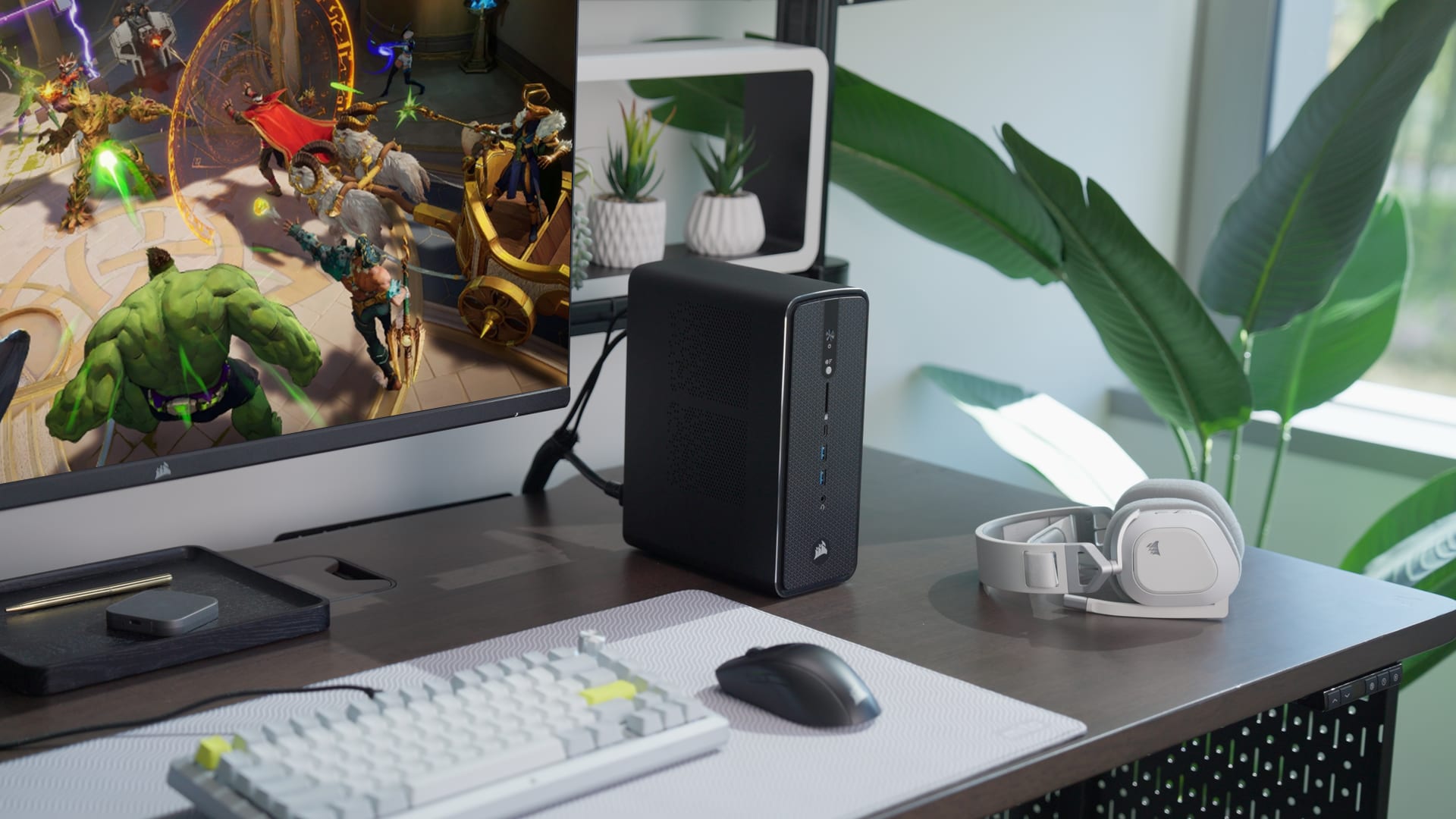 A compact black Corsair AI Workstation 300 desktop PC on a wooden desk alongside a white keyboard, mouse, white Corsair headset on a stand, potted plants, and a monitor displaying a colorful game scene